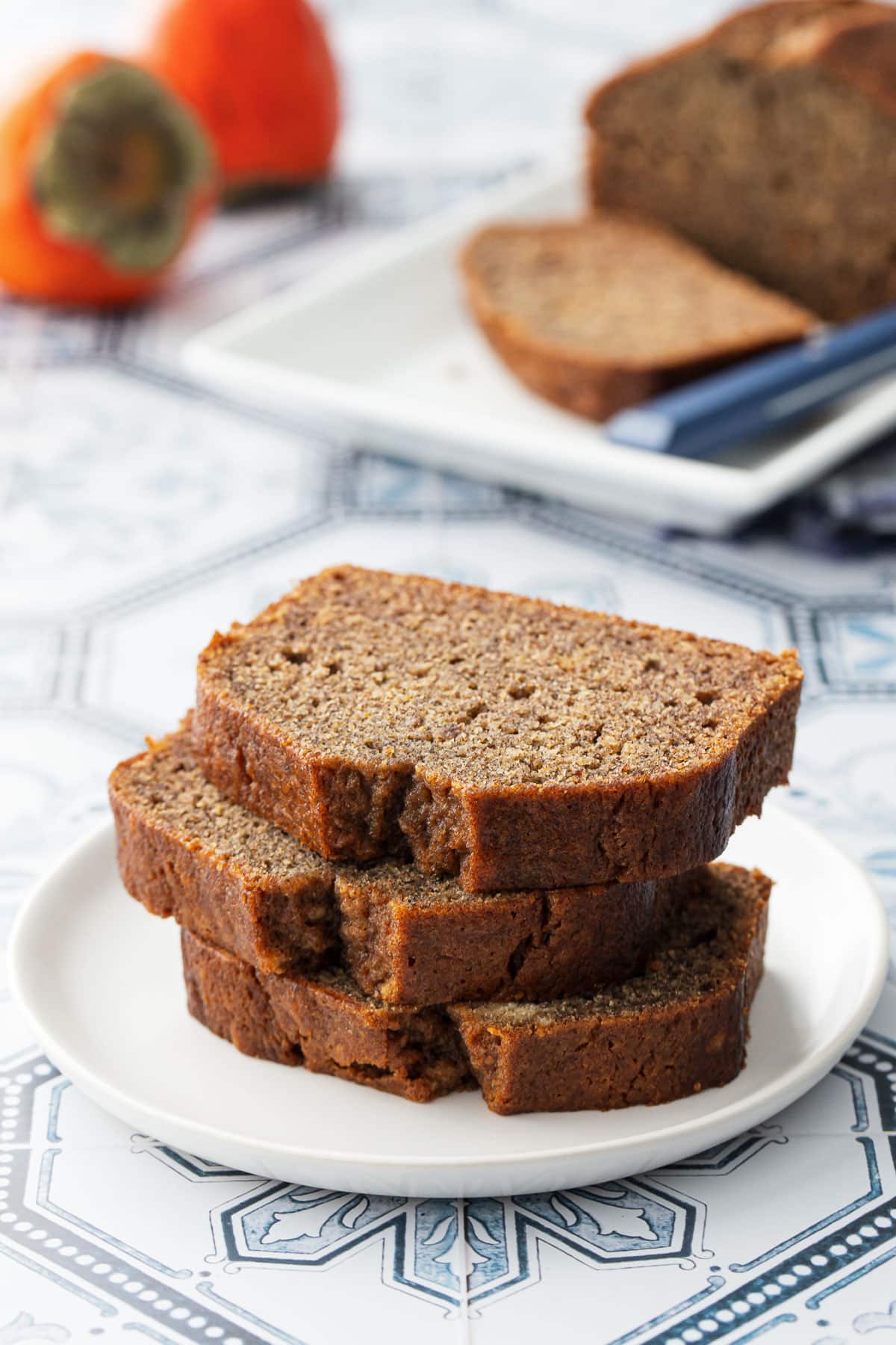 Three slices of Spiced Persimmon Bread stacked on a white plate, with the rest of the loaf and two persimmons in the background.