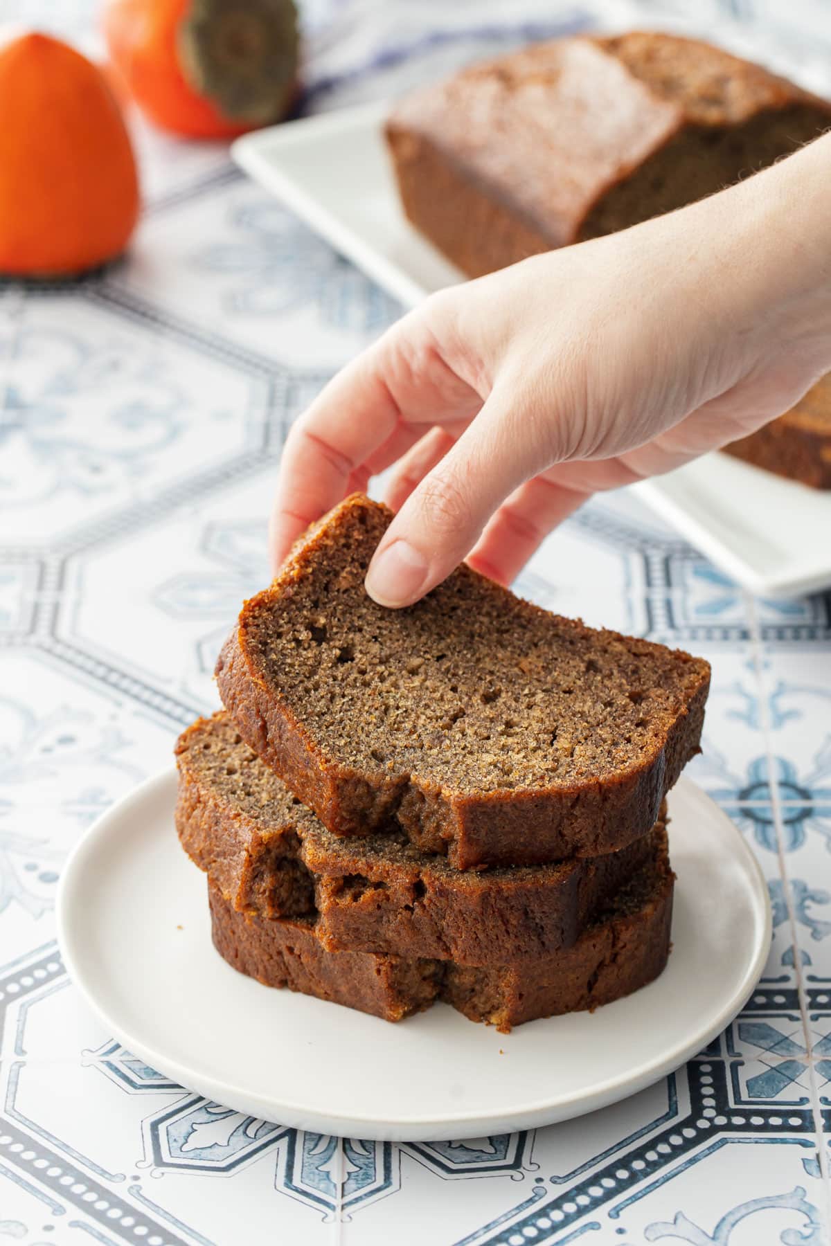 Slices of Spiced Persimmon Bread stacked on a white plate, hand lifting the top slice to show the moist texture.