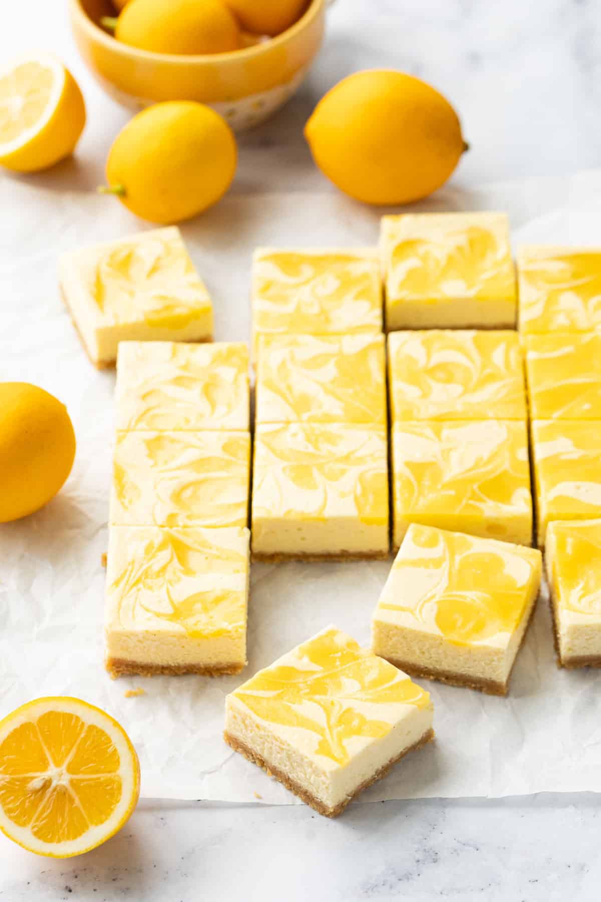 Squares of Meyer Lemon Cheesecake Bars with a swirl of lemon curd on top, on a piece of parchment with a few Meyer lemons scattered around.