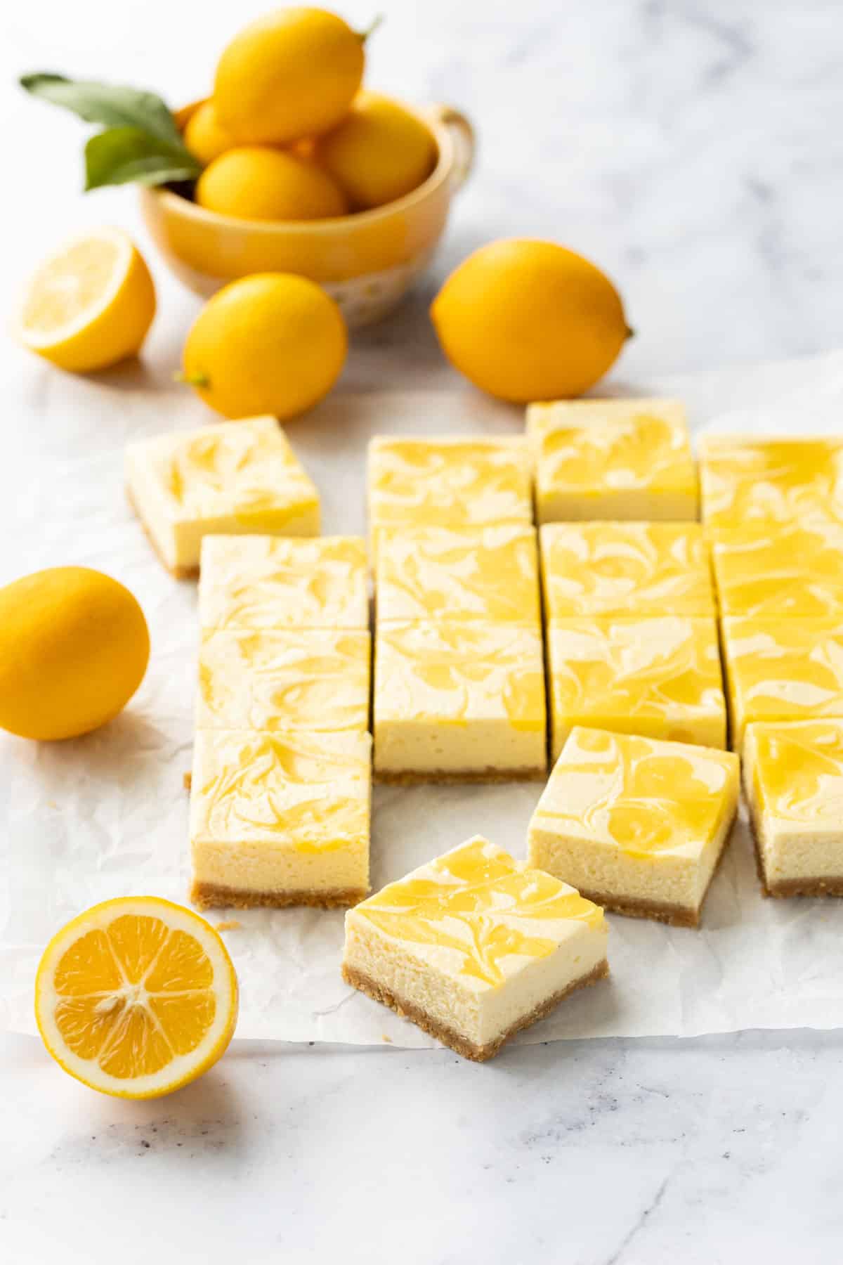 Cut squares of Meyer Lemon Cheesecake Bars with a swirl of lemon curd on top, on a piece of parchment with a few cut and whole Meyer lemons and a bowl of lemons in the background.