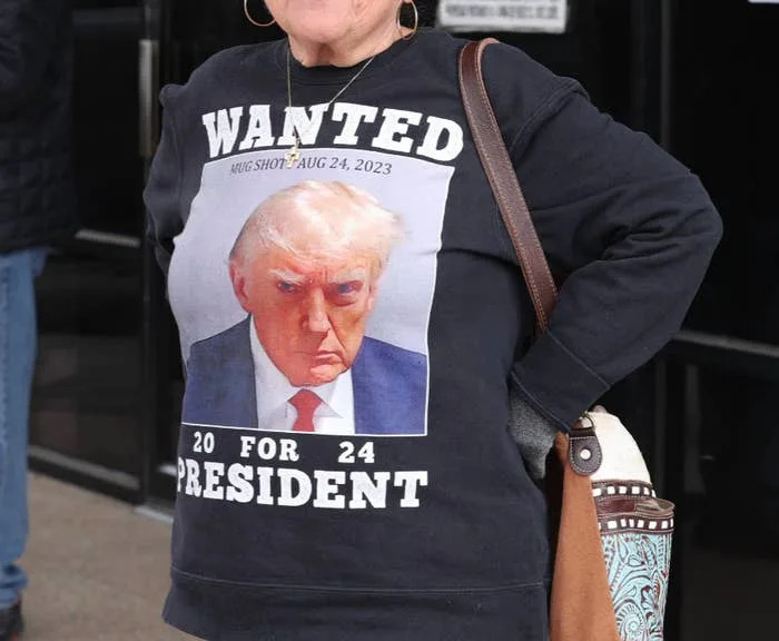 A woman wearing a "WANTED: President" sweatshirt with a photo of a public figure, standing outside a building with visible text signage