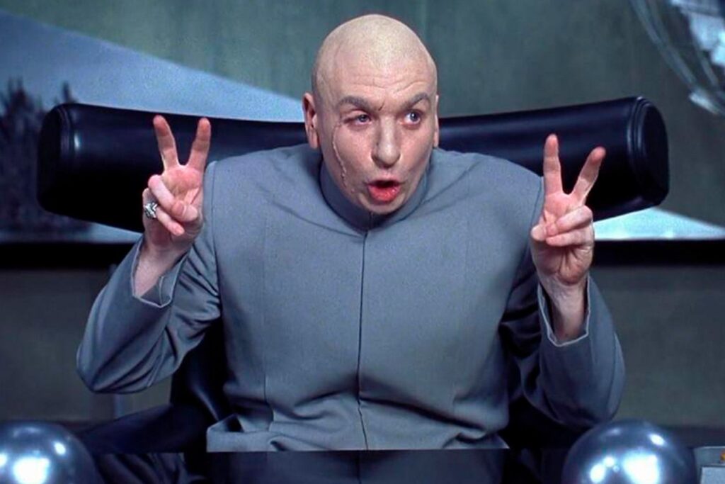 A bald man in a gray suit sits in a black chair, raising both hands and making air quotes with his fingers. He has a serious expression and a noticeable facial scar.