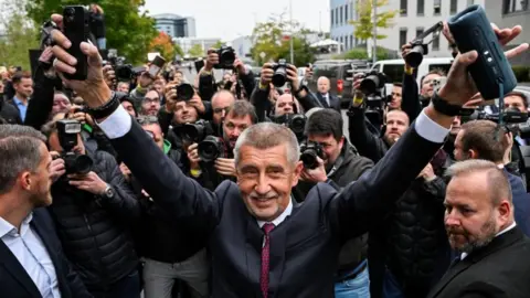 Reuters Andrej Babis dressed in a dark suit with his hands in the air in celebration, surrounded by journalists taking photos of him.