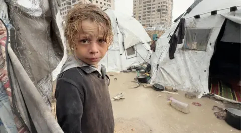 BBC A Palestinian child, wet from rain, looks into the camera, surrounded by soaked tents and wet and muddy ground.