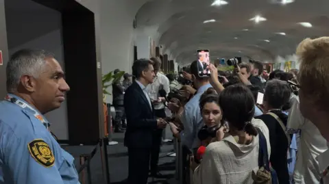 Tom Ingham/BBC Mr Miliband stands wearing a suit stands behind a security line with a crowd of journalists.