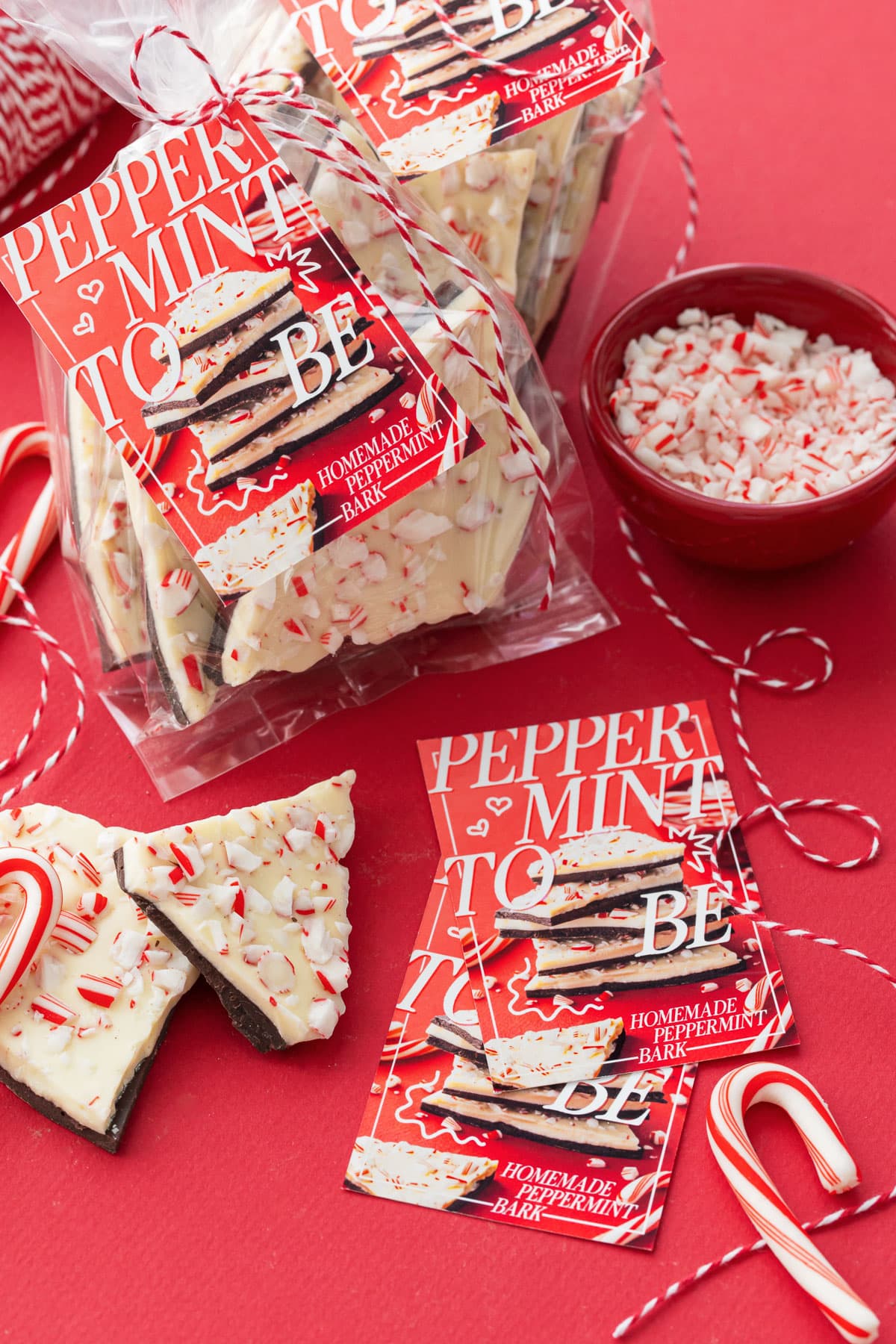 Clear treat bag filled with Homemade Peppermint Bark on a red background, tied with twine and printable gift tags, a few more tags and a small bowl of crushed candy canes off to the side.