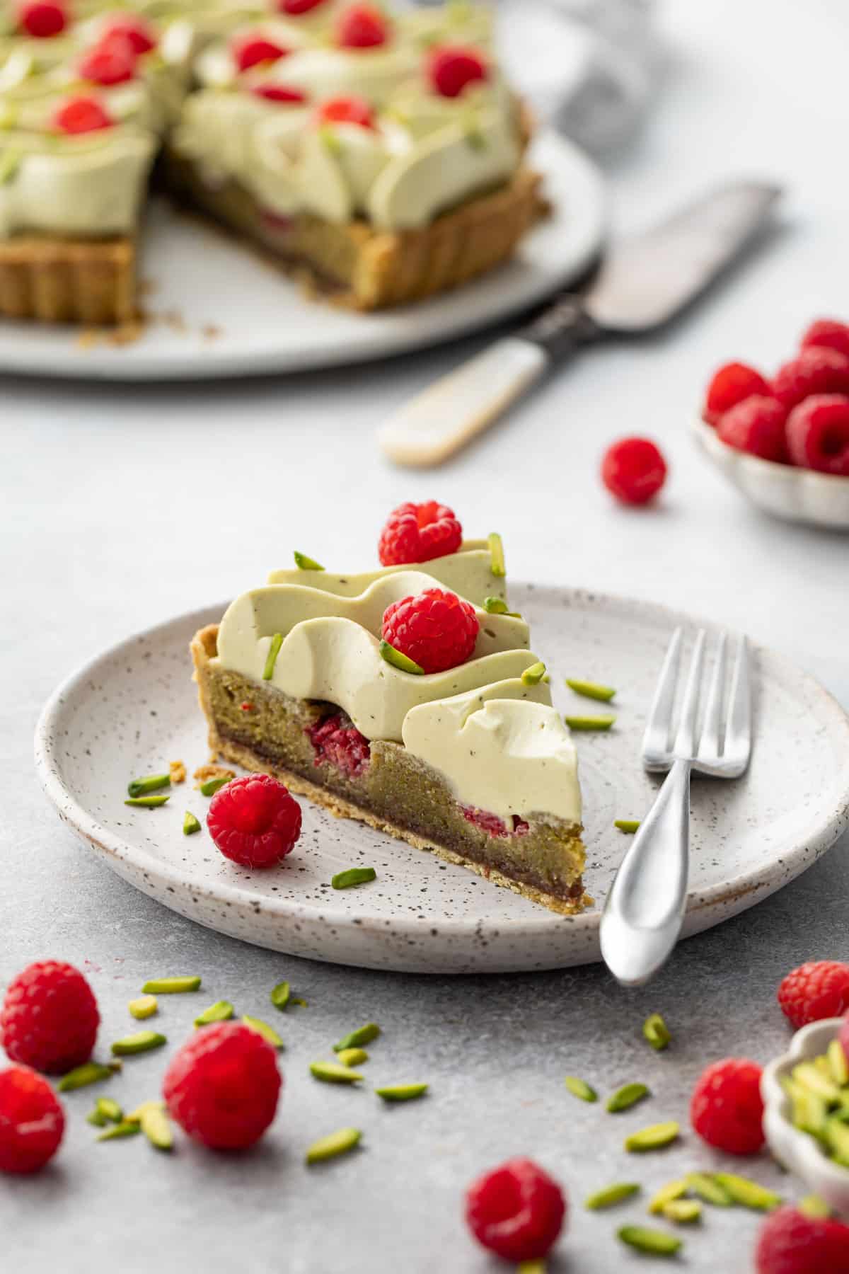 Slice of Raspberry & Pistachio Frangipane Tart with Whipped Pistachio Ganache on a speckled plate, with a fork and the rest of the tart in the background.